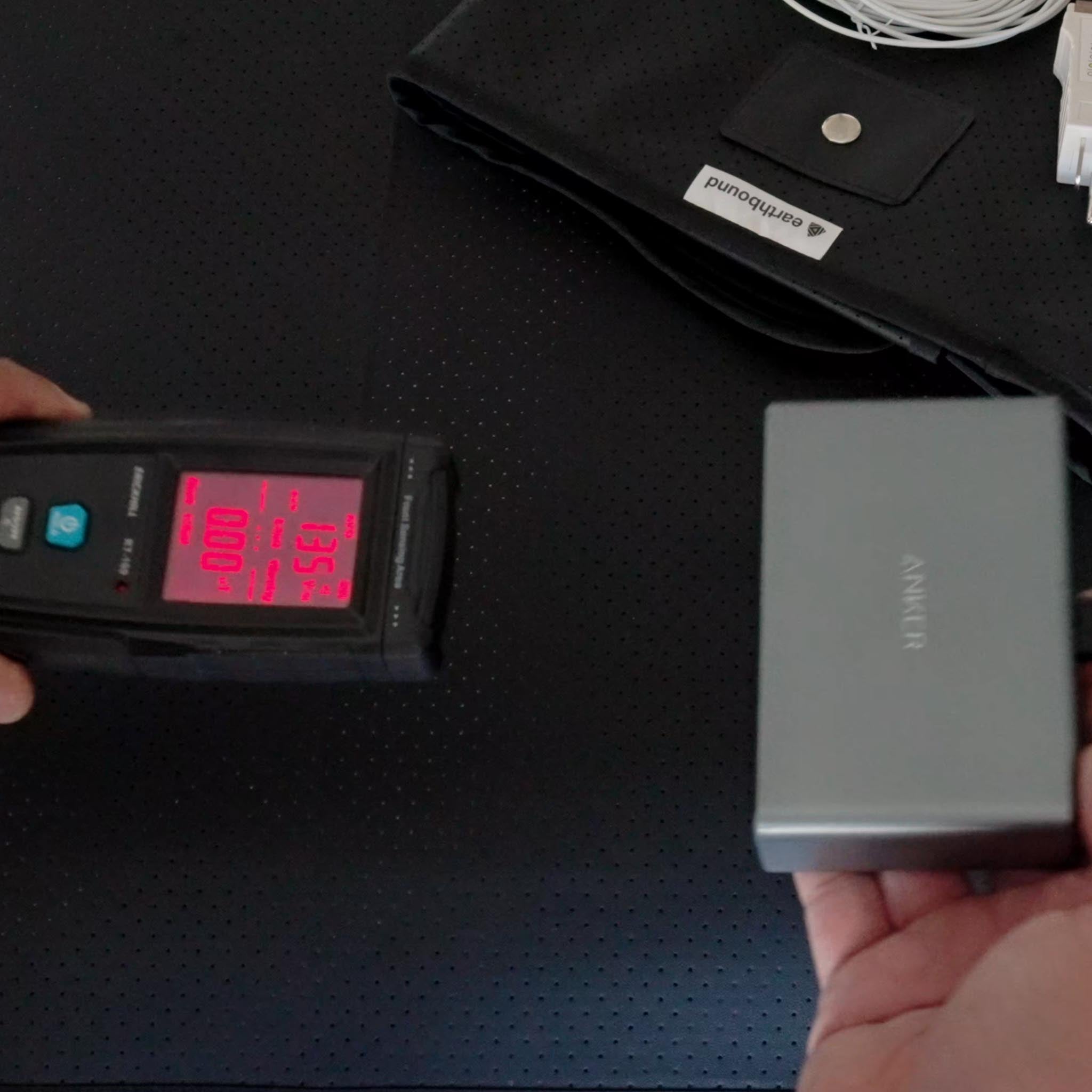 EMF meter measuring radiation levels near grounding mat and power bank showing 100 reading on display
