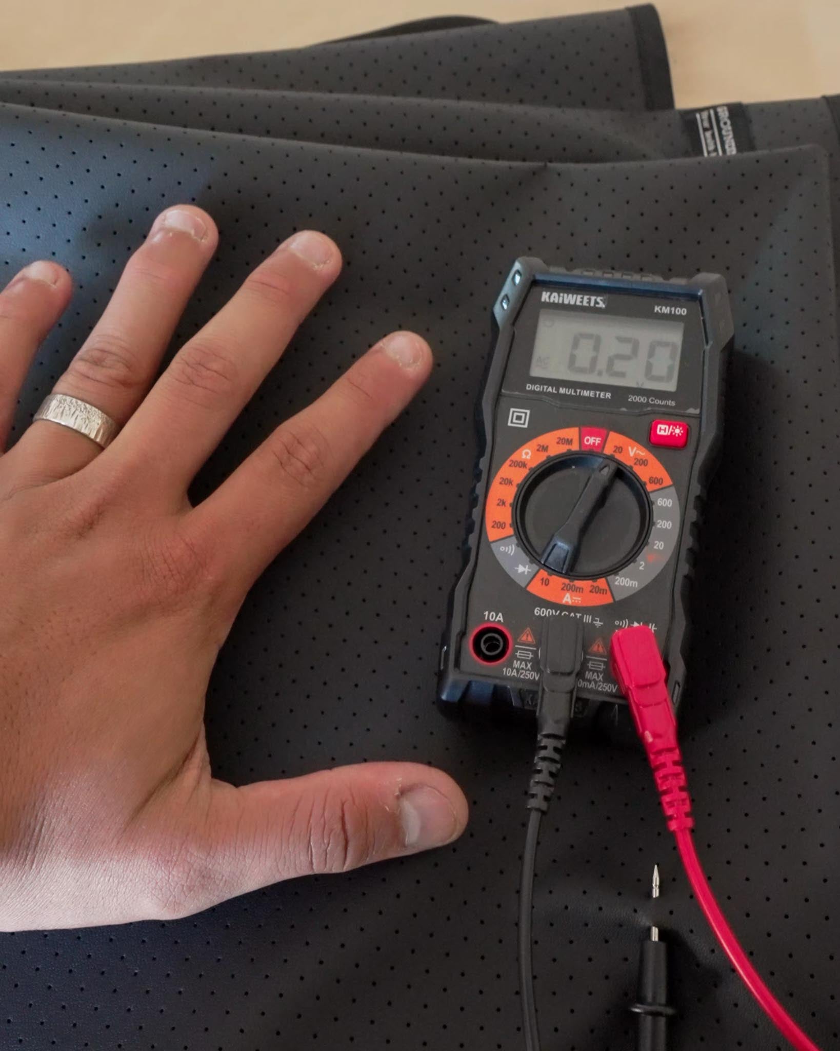 Hand on grounding sheet showing low voltage reading (0.20V) on multimeter